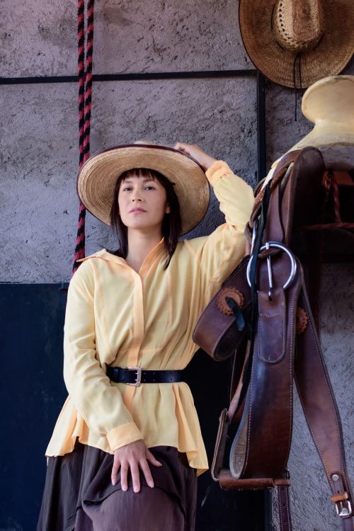Free Hispanic woman posing fashionably in a yellow blouse and brown hat beside equestrian equipment indoors. Stock Photo