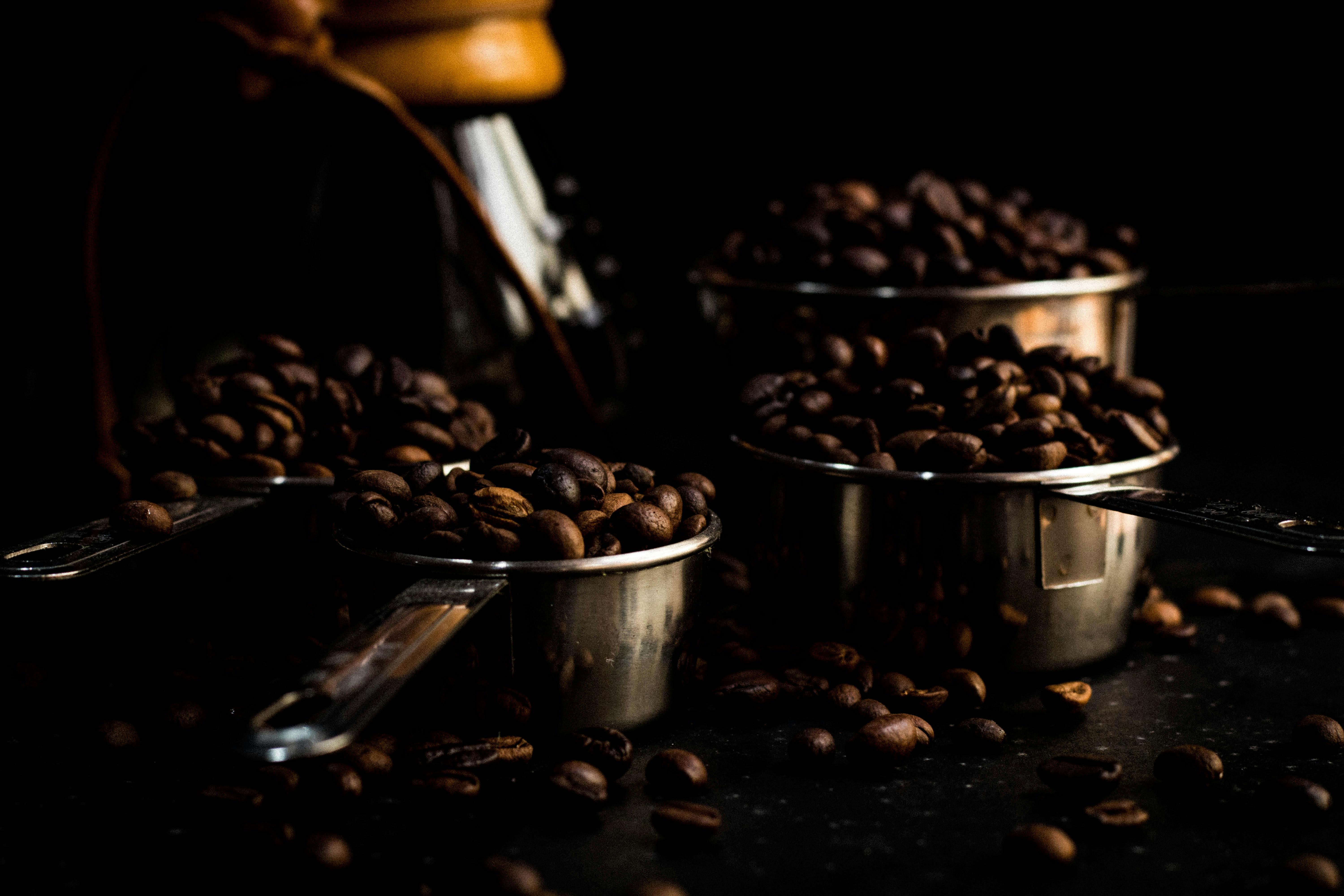 Roasted coffee beans overflowing from metal measuring cups in low light setting.