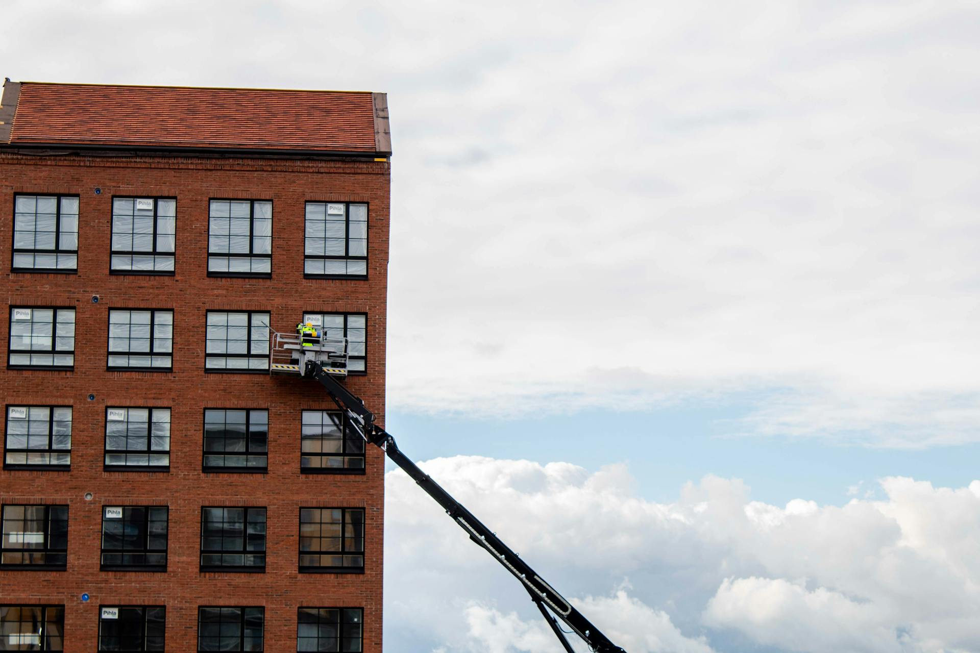 Worker on a telescopic lift inspecting a red brick building against a cloudy sky.