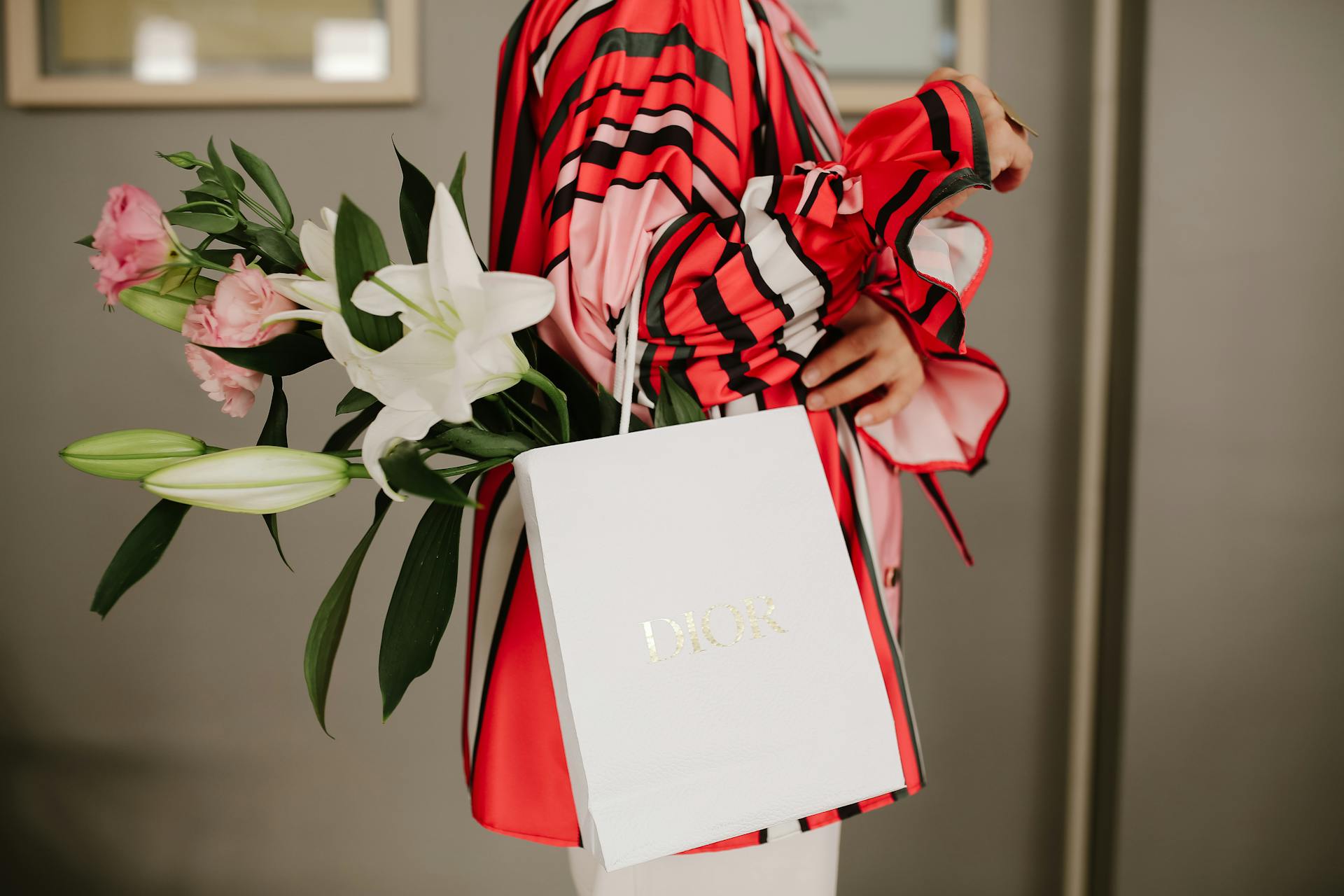 Stylish woman in patterned blouse holding bouquet and gift bag indoors.