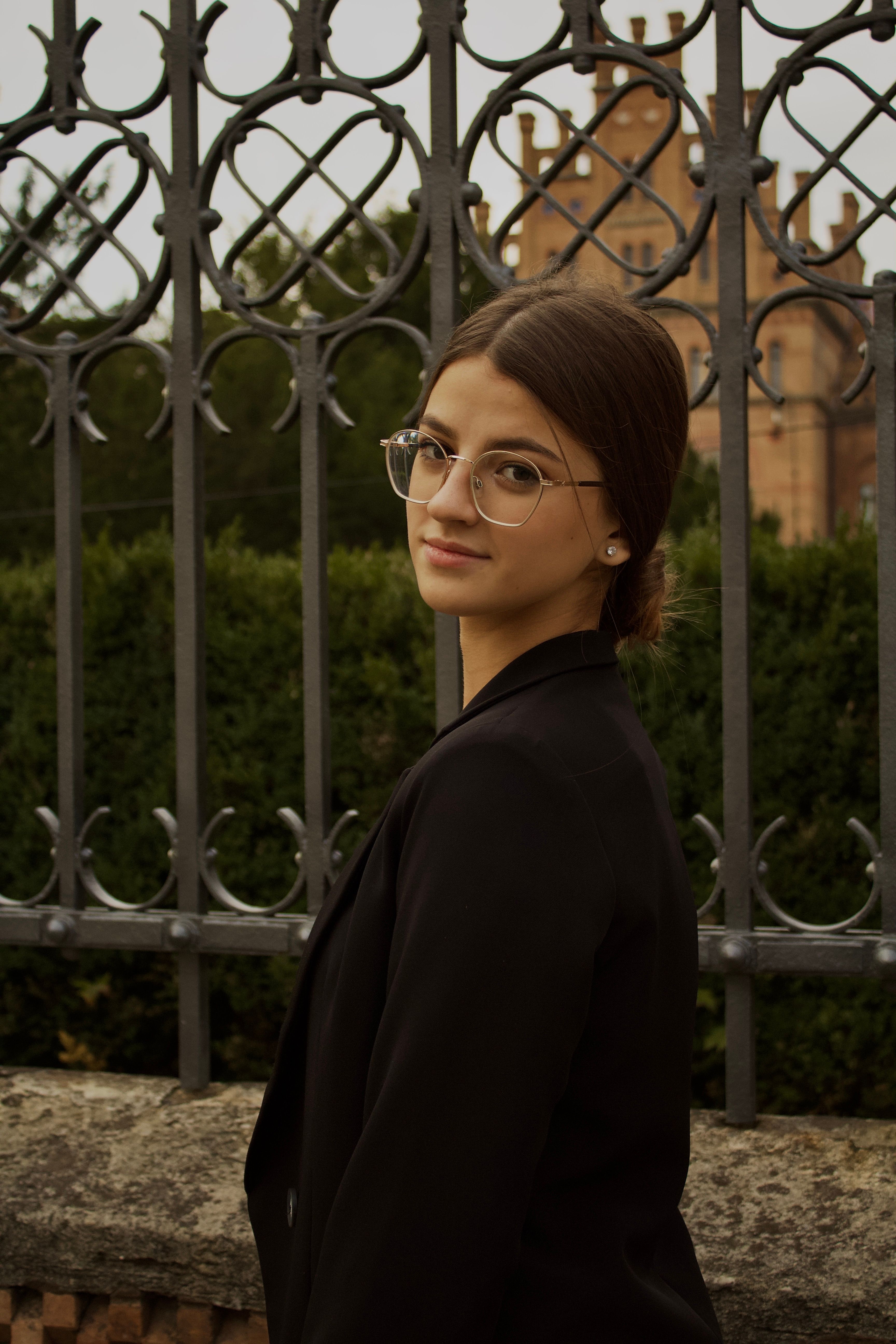 Portrait of a young woman wearing eyeglasses near an ornate fence outdoors.