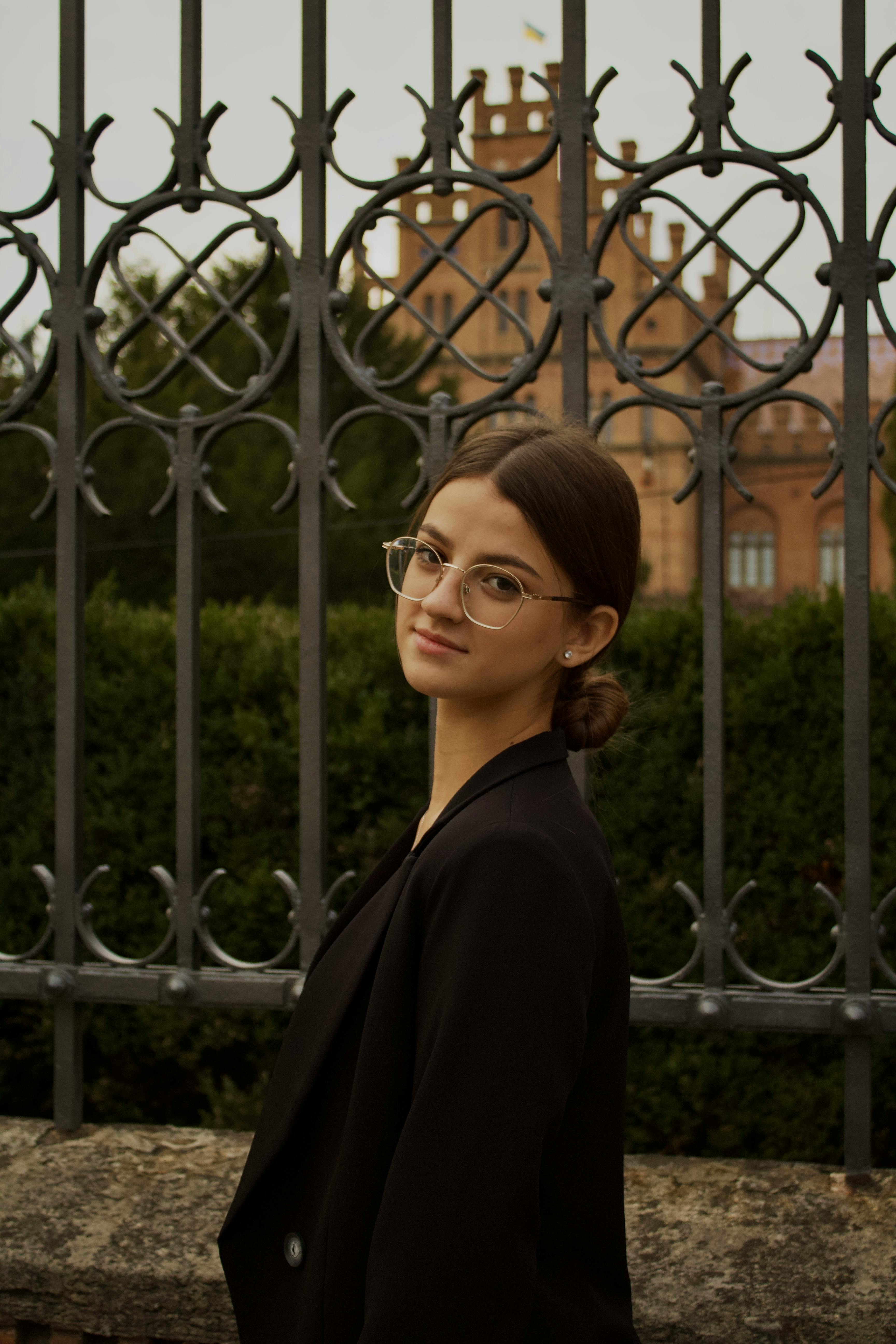 A sophisticated young woman in formal attire stands gracefully before an ornate iron fence.