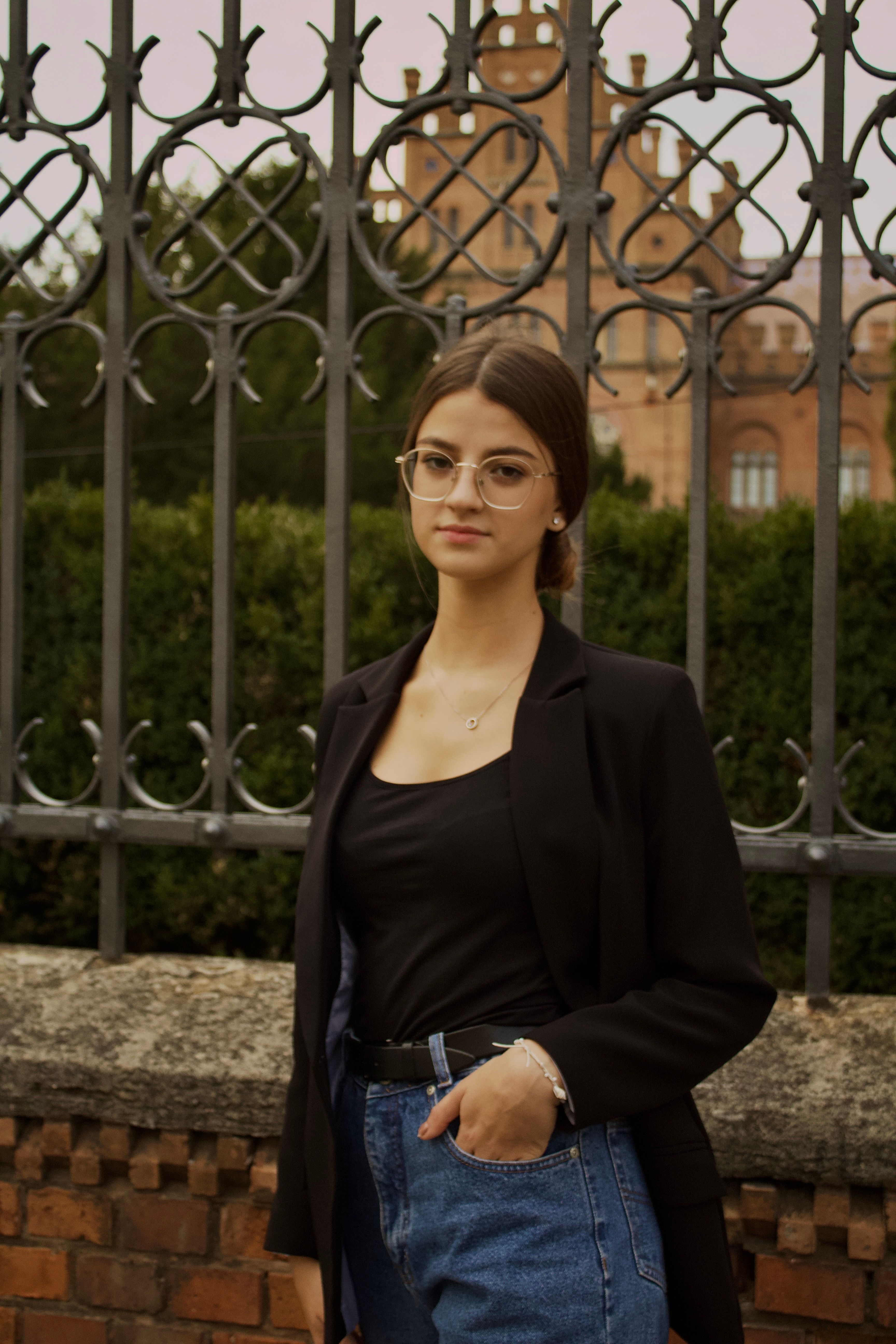 Young woman in elegant black blazer poses outdoors against a decorative fence, exuding a modern fashion sense.
