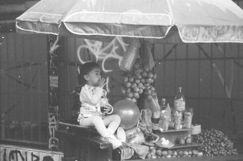Grayscale Photo of Child Sitting on Fruit Stand