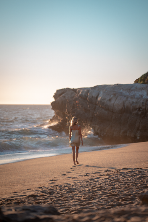 Free A woman in a dress walks along a sandy beach near rocky cliffs during sunset. Stock Photo