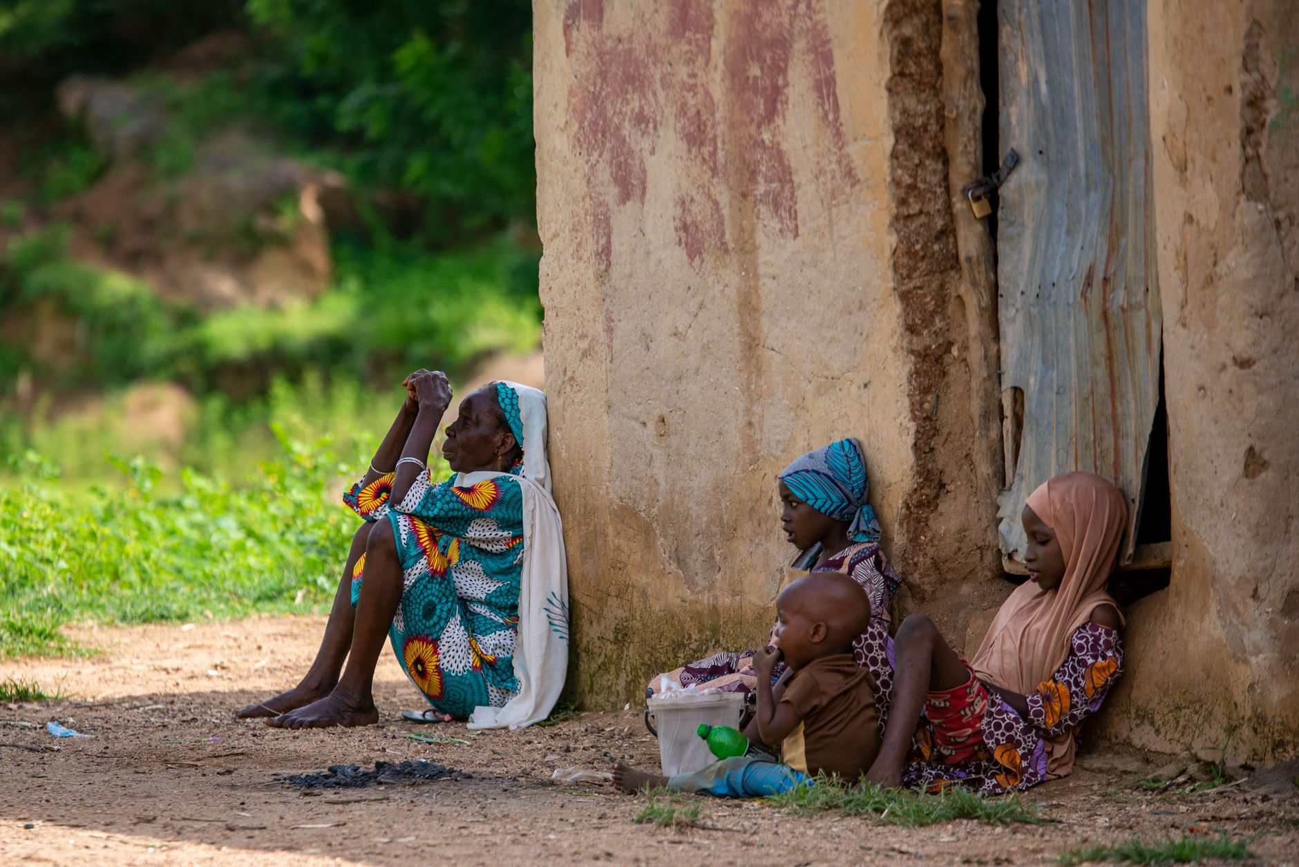 A family rests by a weathered wall in Kano, Nigeria, capturing a moment of daily life.