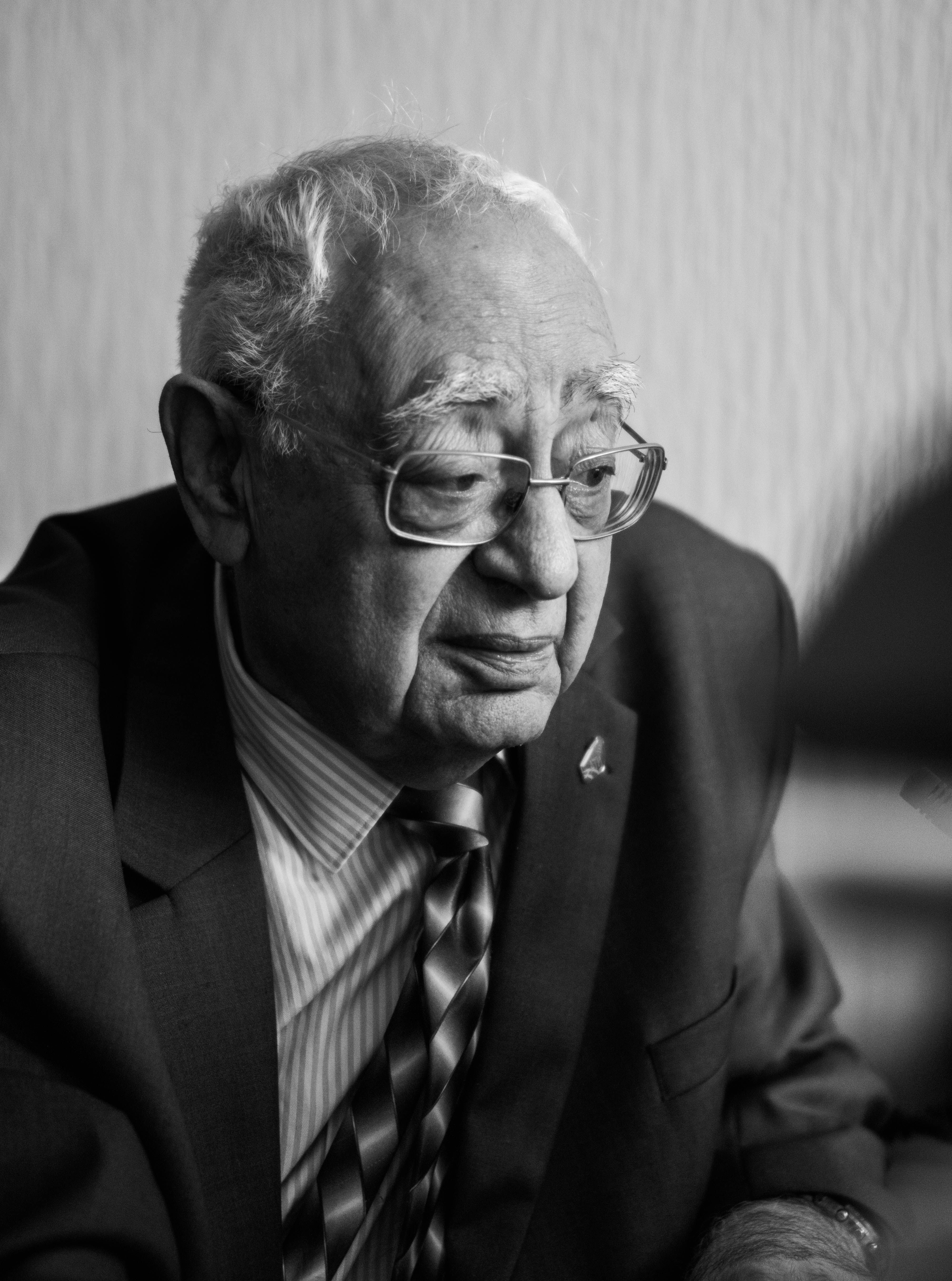 Black and white portrait of a senior man in a suit with glasses, looking contemplative.