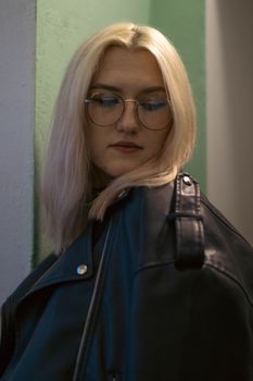 A stylish woman with blonde hair and eyeglasses in a black leather jacket looks down in an indoor setting.