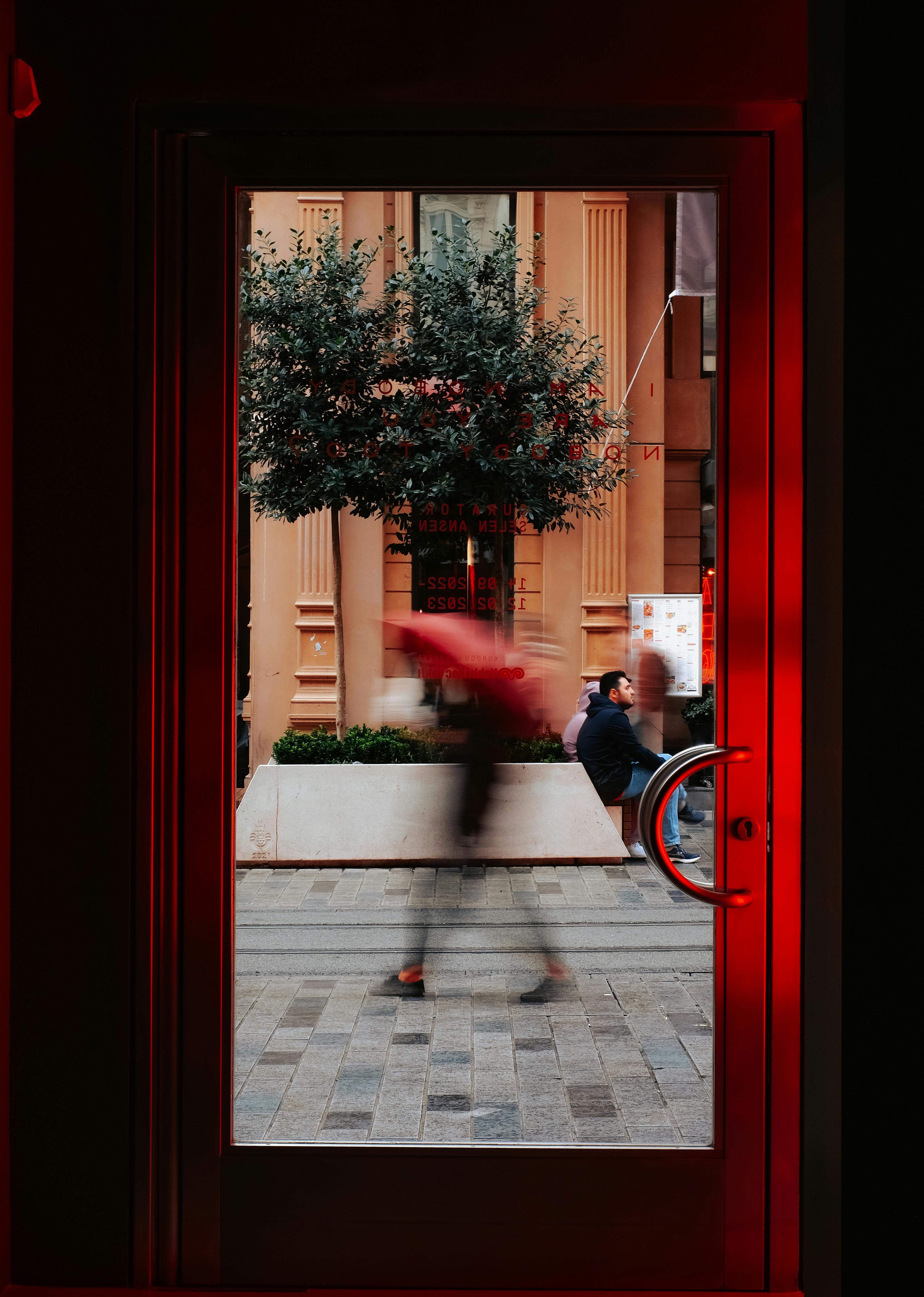 A blurred person with red umbrella captured through a glass door, creating a dynamic urban scene.