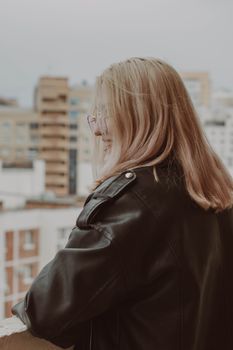 A young woman in a leather jacket enjoys a city view from a balcony, capturing a serene outdoor moment.