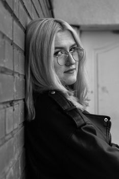 A black and white portrait of a woman leaning against a brick wall, wearing a leather jacket and eyeglasses.