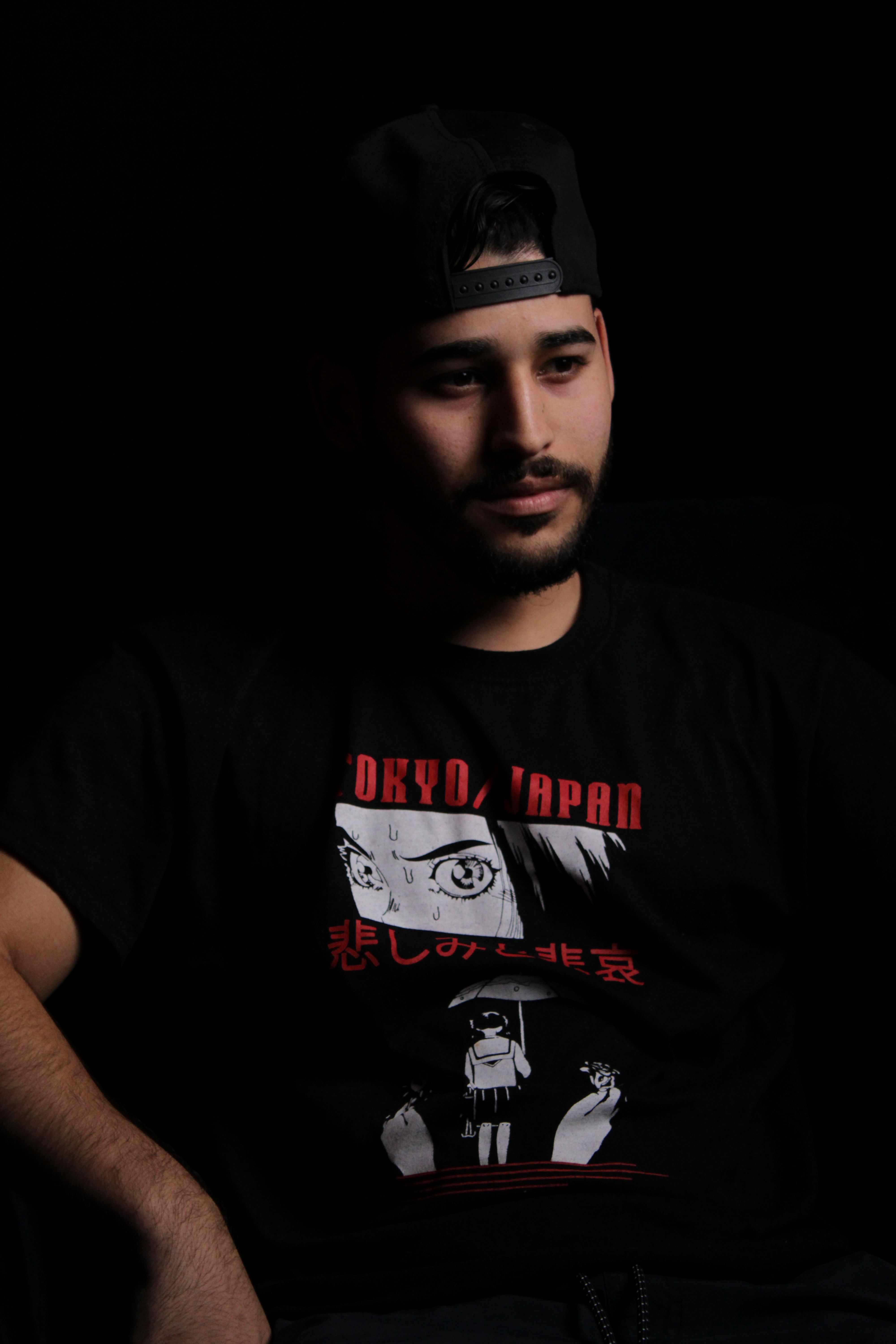 Portrait of a bearded man in a graphic t-shirt and cap, against a dark studio background.