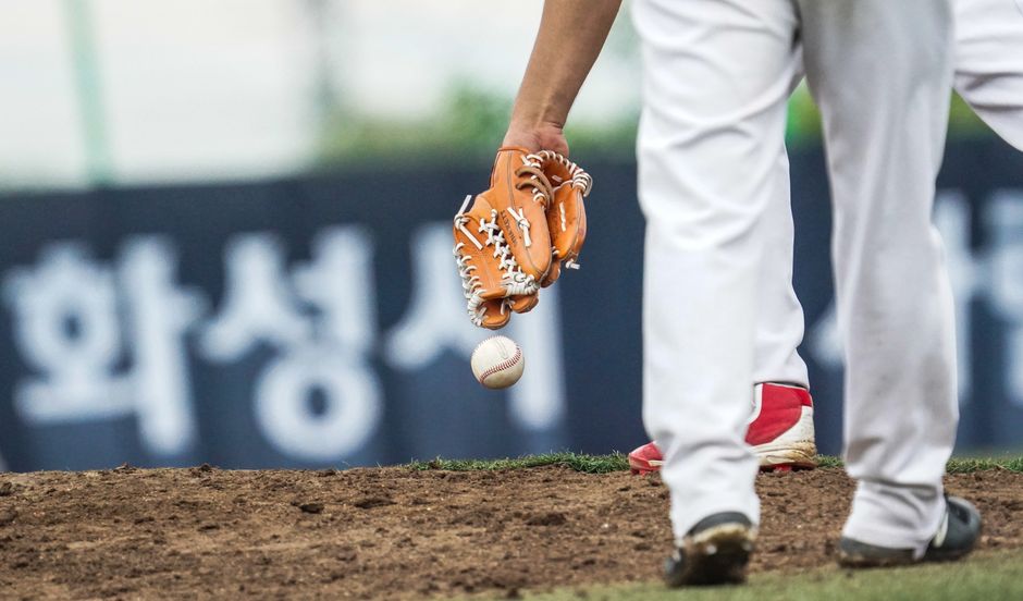 Baseball glove and ball on the infield dirt of a Korean ballpark, with Hwaseong city (화성시) signage visible on the outfield wall — Photo by 준섭 윤 on Pexels