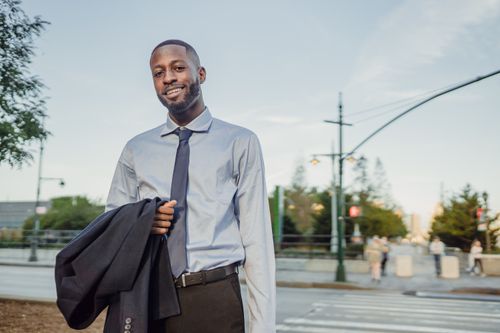 Free Businessman in formal attire smiling and holding a suit jacket outdoors in an urban setting. Stock Photo