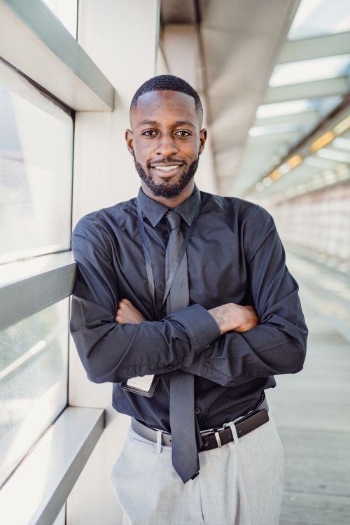 Free Businessman in a sleek black shirt standing confidently with arms crossed in a contemporary office hallway. Stock Photo