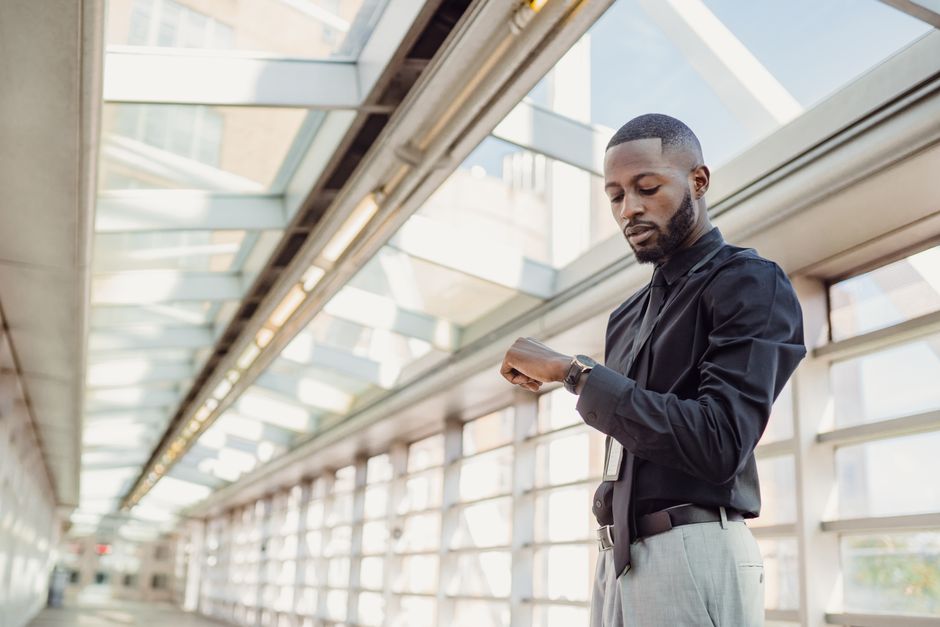African American man in shirt checks wristwatch, standing in vibrant modern glass corridor. African American man in shirt checks wristwatch, standing in vibrant modern glass corridor.