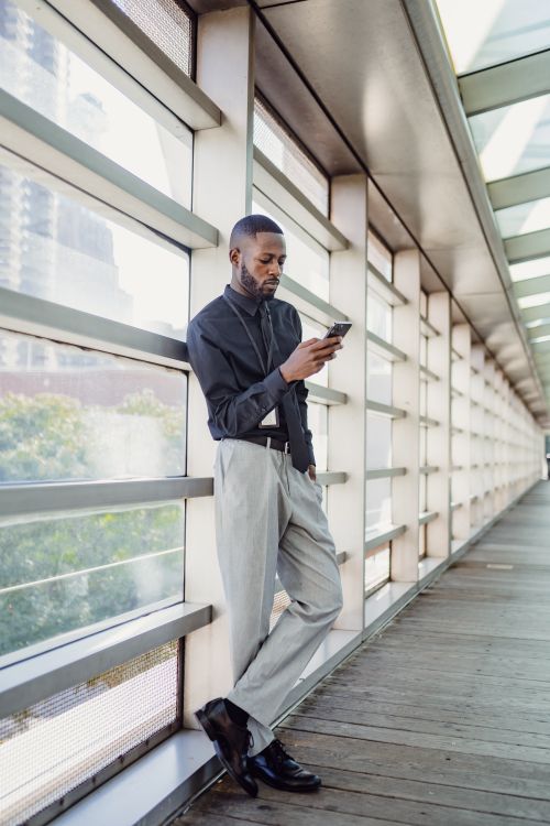 Free African American man leans against a glass wall, engaging with his smartphone in a modern urban environment. Stock Photo