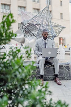 African American businessman using laptop near modern sculpture, showcasing urban work lifestyle.
