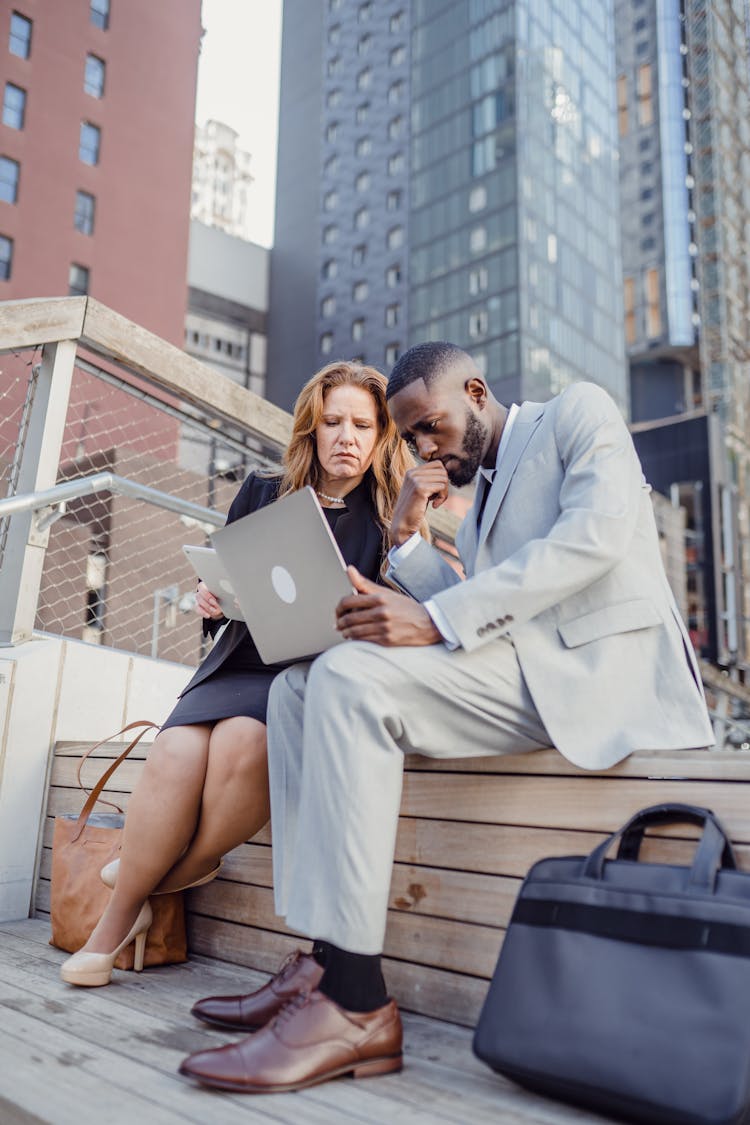 Businessman And Businesswoman Sitting On A Wooden Step In A City And Looking At Laptop