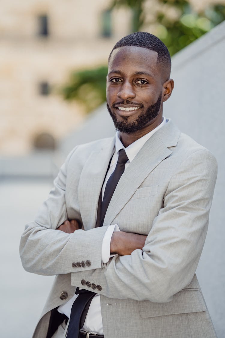 Smiling Man In A Gray Suit Standing With Arms Crossed By A Fence