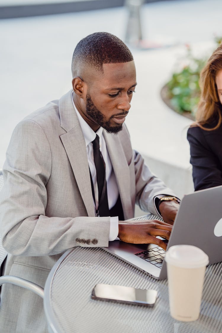 Man In A Gray Suit Sitting At A Metal Table And Typing On A Laptop