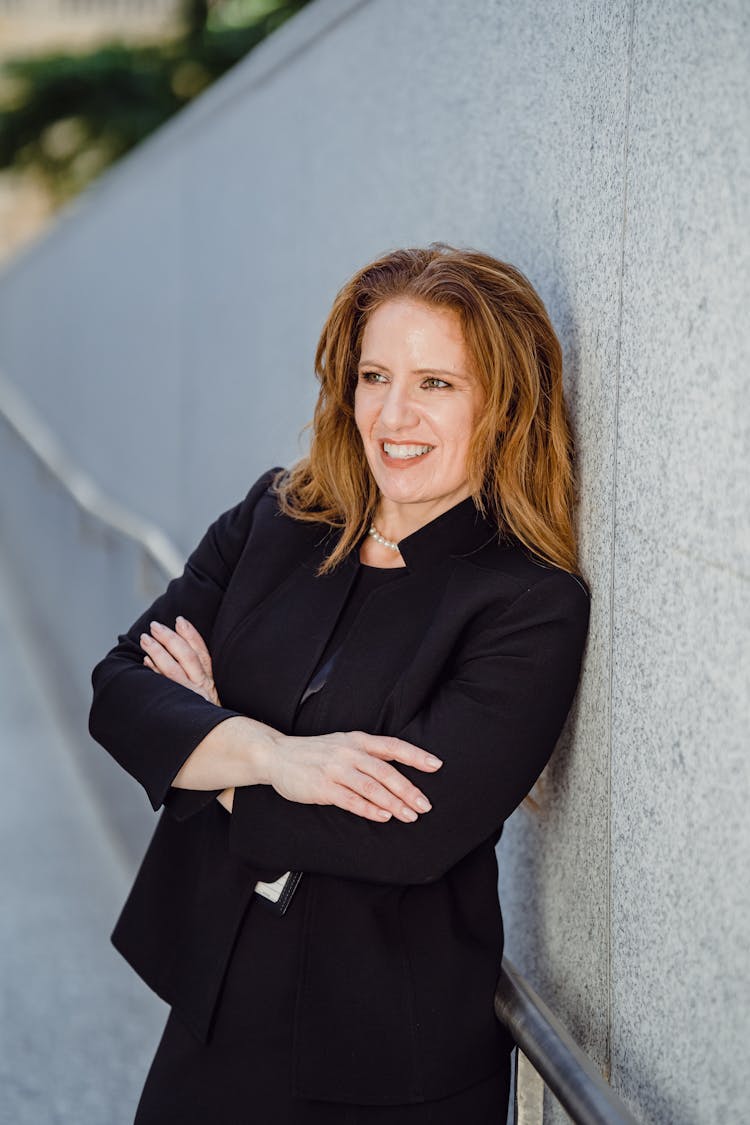 Woman Wearing A Black Suit Leaning Against A Marble Wall With Arms Crossed And Smiles