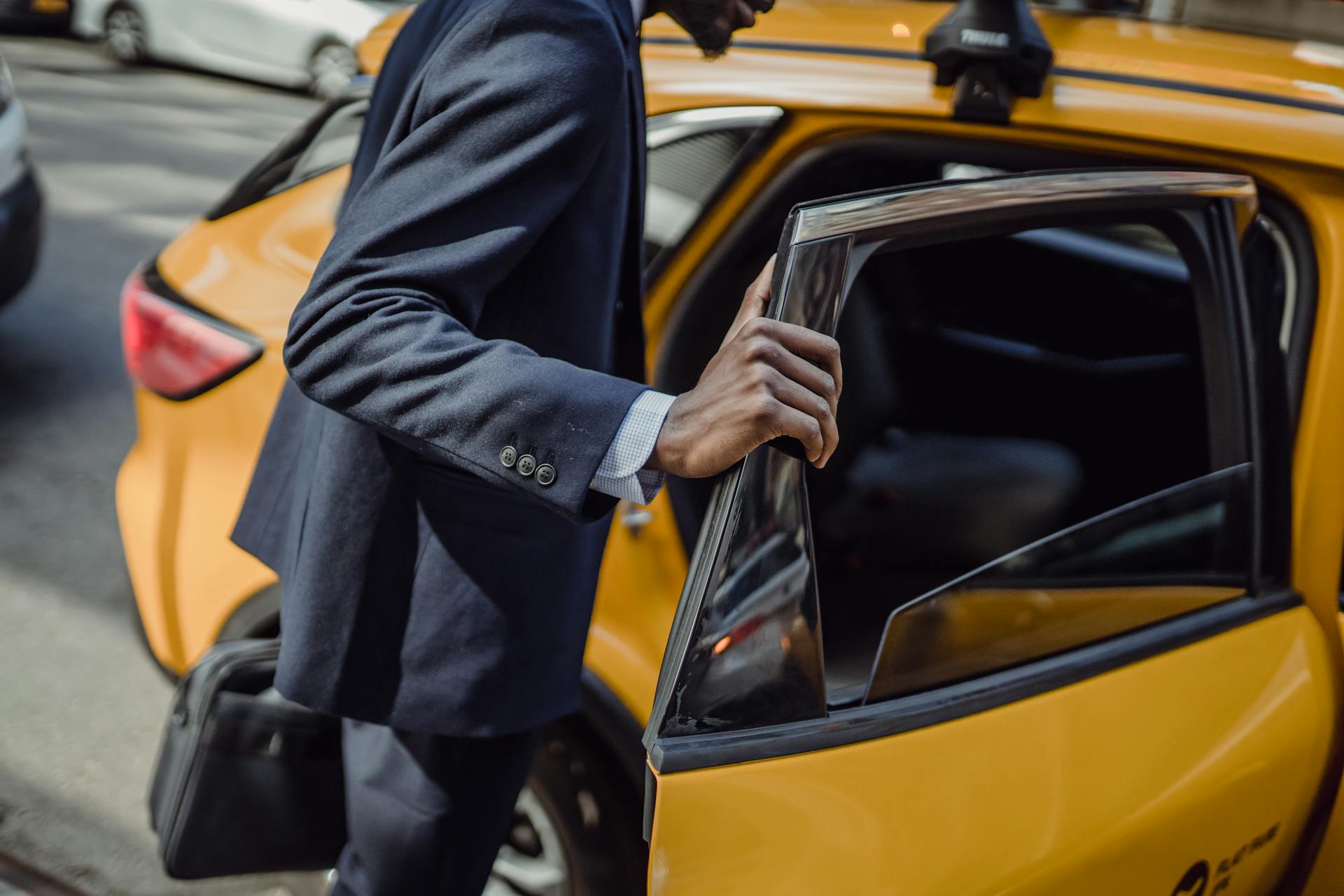 A businessman in a suit opens the door to a yellow taxi on a city street during the day.