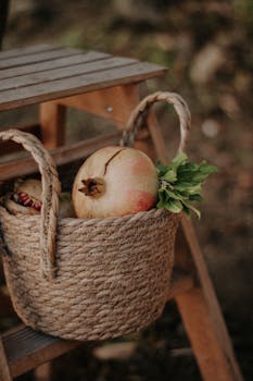 A rustic woven basket holding fresh pomegranates sits on a wooden stool in an outdoor setting.