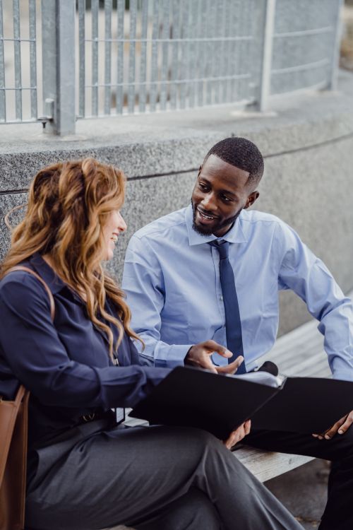 Free Two professionals sit on a bench discussing documents in a business setting outdoors. Stock Photo