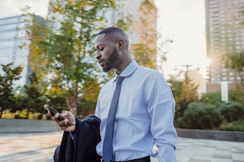 Free A young professional using his smartphone amidst tall city buildings and trees. Stock Photo