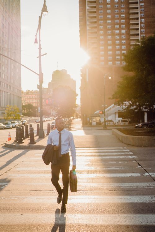 Free A man in professional attire walks across a city street during sunset, carrying a bag. Stock Photo