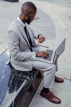 A professional man in a suit checks his wristwatch while using a laptop outdoors, portraying modern business.