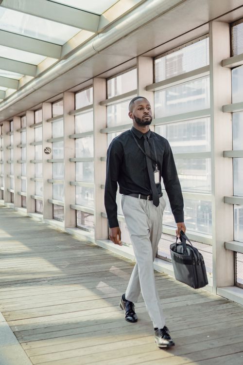 Free Confident business professional walking in a glass-covered hallway holding a laptop bag. Stock Photo