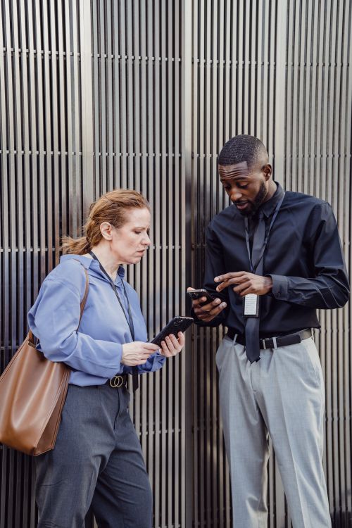 Free Two professionals in business attire discuss work outside an office, using smartphones. Stock Photo
