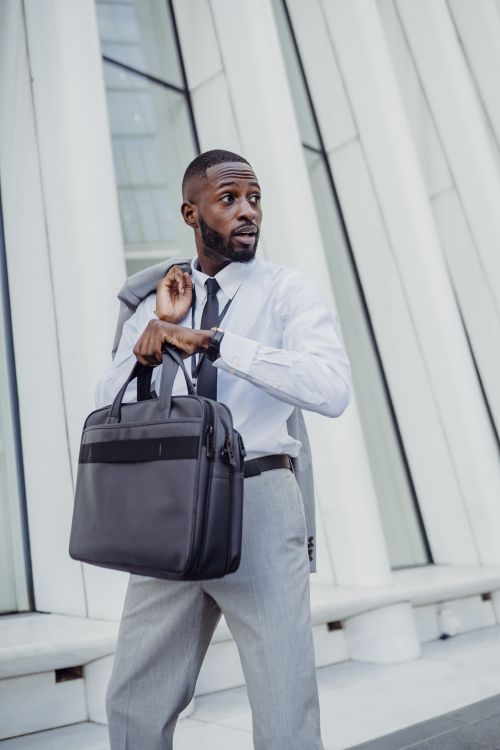 Free A businessman in formal attire holding a laptop bag outside a contemporary building, checking the time. Stock Photo