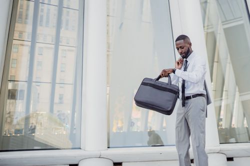 Free Professional man checking time with wristwatch while holding a laptop bag outside modern office. Stock Photo