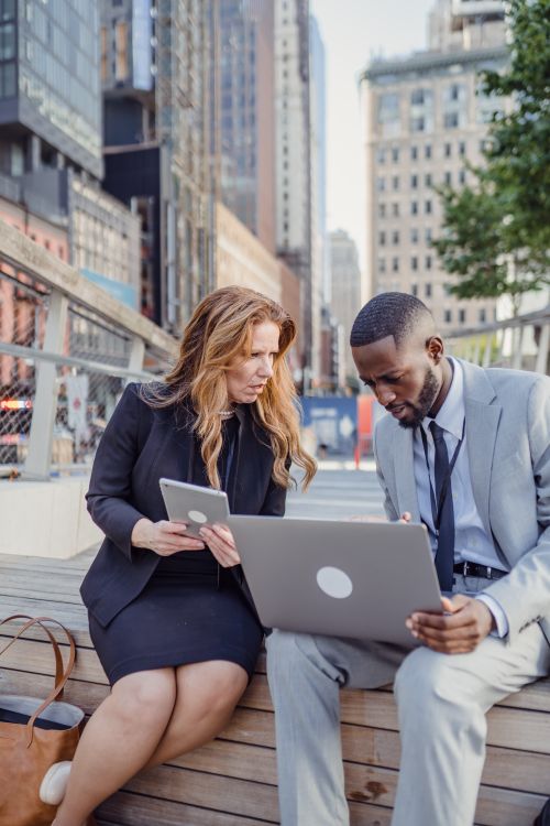 Free Two business professionals discuss work on a laptop and tablet outdoors in an urban setting. Stock Photo