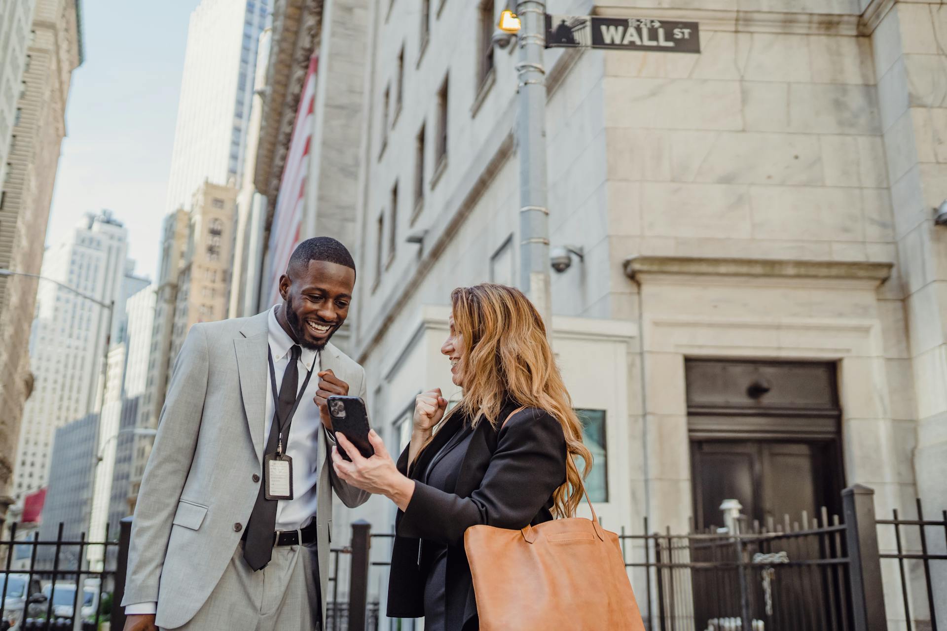 Two business professionals in suits smiling and viewing a cellphone outdoors on Wall Street.