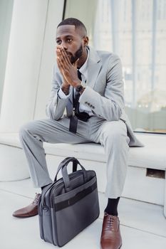 A thoughtful man in a suit sits indoors with a briefcase on a bench, suggesting introspection and business focus.