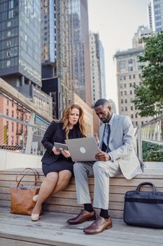 Two business professionals on a city bench working on a laptop.