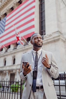 Smiling businessman in a suit stands in front of a stock exchange building, holding a cellphone.