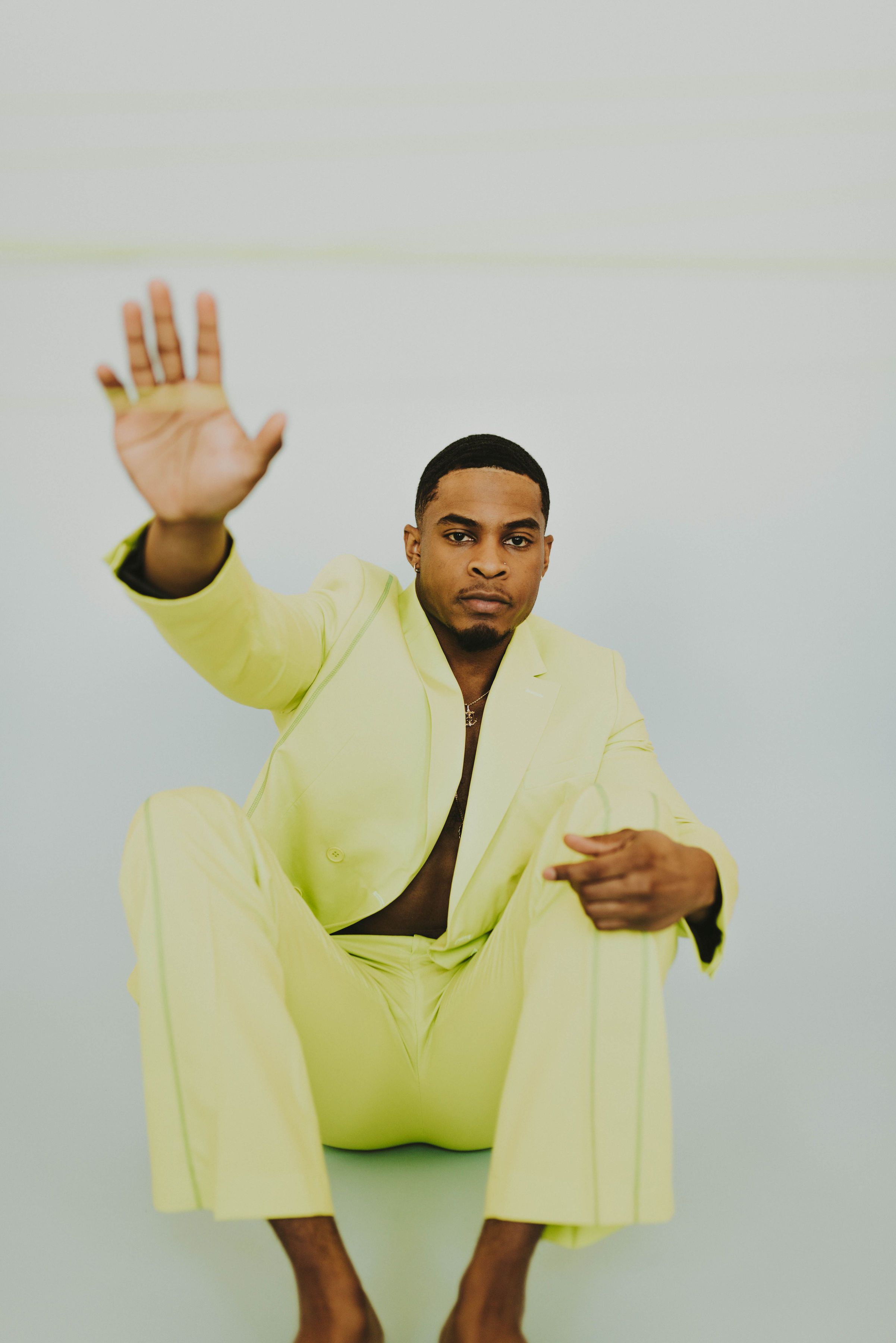 Fashion-forward sitting pose with arm raised in a vibrant neon suit against a white background.