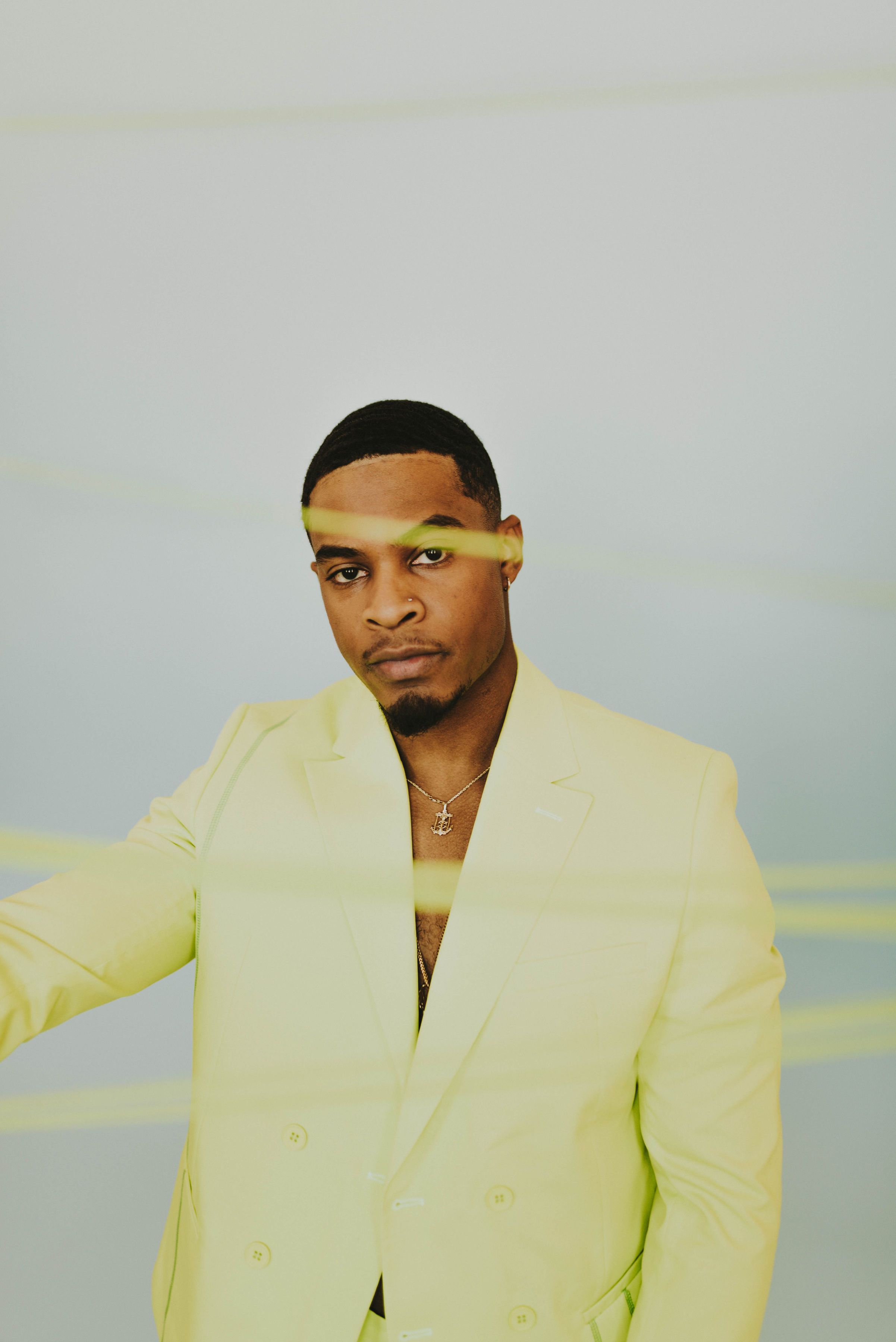 Fashionable black male model posing in vibrant yellow blazer against gray background with necklace.