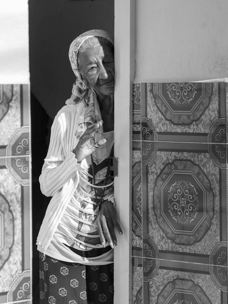 Grayscale Photo Of An Elderly Woman Hiding Behind The Wall