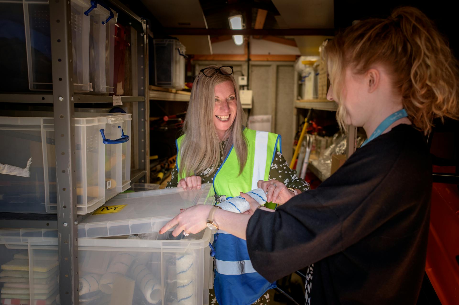 Two women in a warehouse organizing containers with smiles, promoting teamwork.