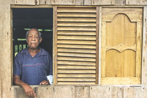 Free Senior man standing in a wooden house window, blue polo shirt, serene expression. Stock Photo