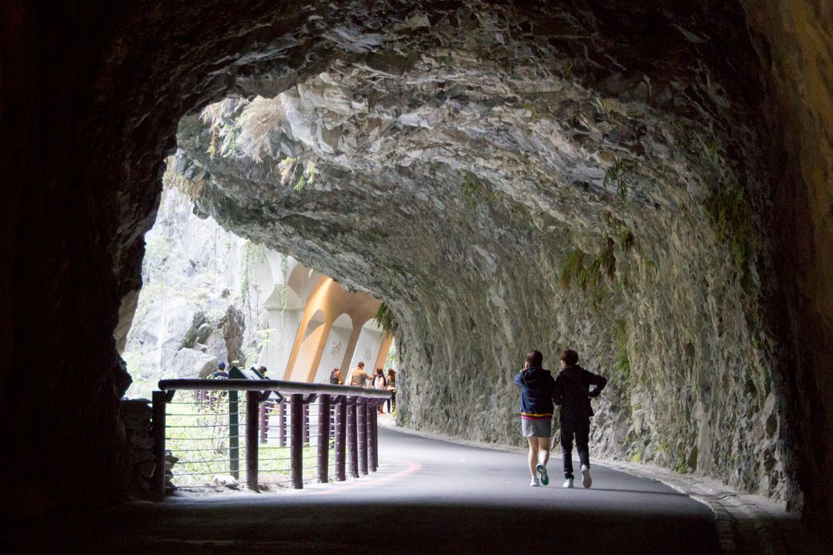 Hikers walking through a marble tunnel carved into the gorge wall, daylight ahead.