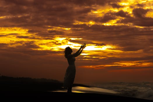A woman stands on the beach with arms raised, celebrating a stunning golden sunset.