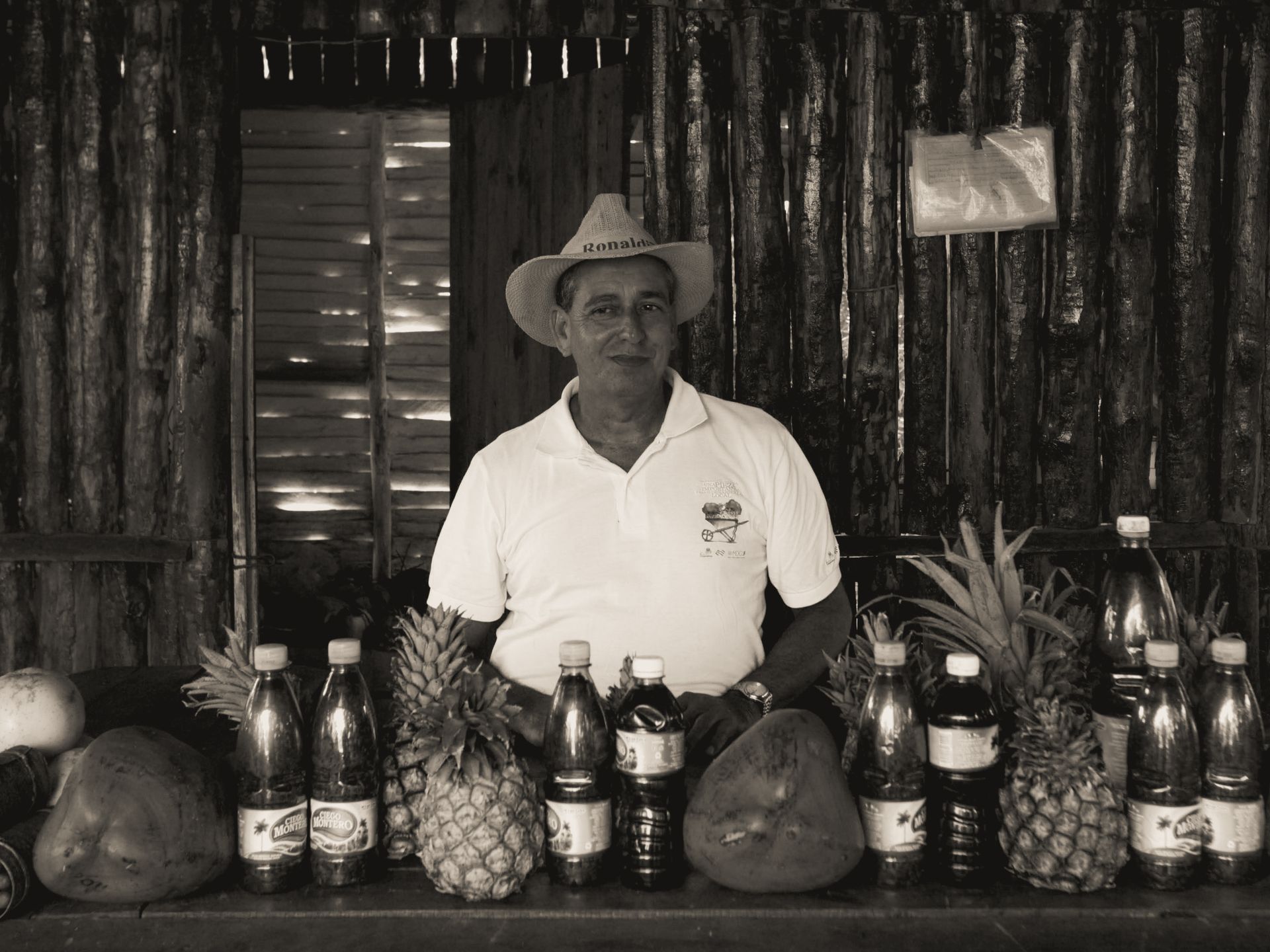 Black and white portrait of a market vendor with fruits and products on display in a rustic setting.