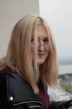 Blonde woman with eyeglasses and leather jacket smiling outdoors.